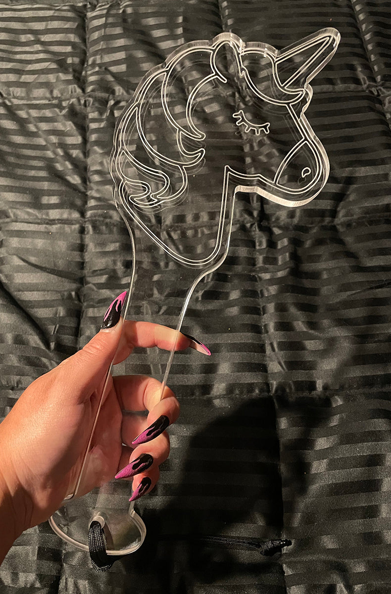 Clear acrylic unicorn-head paddle held in a hand with pink and black nails against a black satin background.
